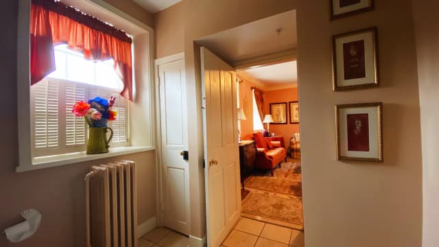 Warm-toned hallway with a side table under a window holding colorful flowers. Open door leads to a cozy living room with a sofa and lamps.