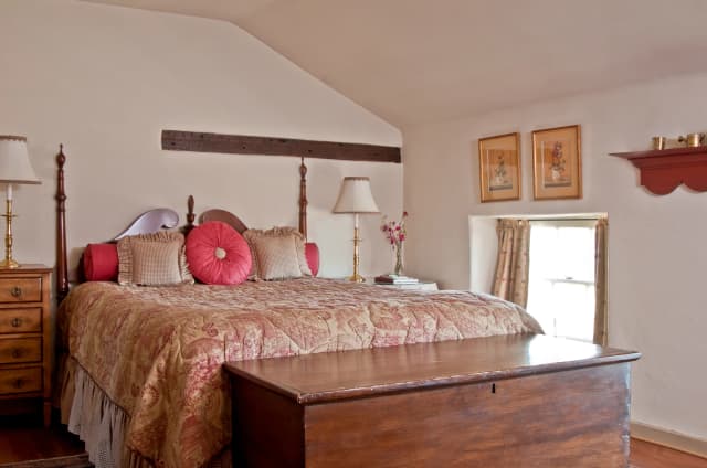 Betty Suite bedroom: Cozy bedroom with a large, ornate bed adorned with red cushions and a floral quilt. Two bedside lamps, wooden chest, and bright window enhance warmth.