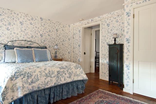 Margo Suite bedroom with floral blue and white wallpaper and bedding. Dark blue dresser, patterned lamps, hardwood floors, and a rug add a vintage touch.
