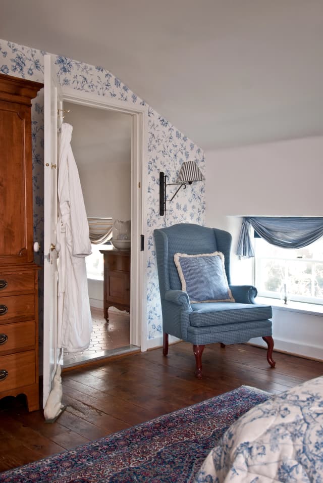 A Margo Suite bedroom corner with floral wallpaper, a blue armchair, and a matching pillow. Warm wooden floors and a window with a blue curtain add to the serene atmosphere.