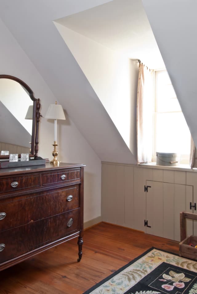 a room with a sloped ceiling, featuring a dark wooden dresser with an ornate mirror, a table lamp, and a window with beige curtains. A floral rug enhances the warm, rustic atmosphere.