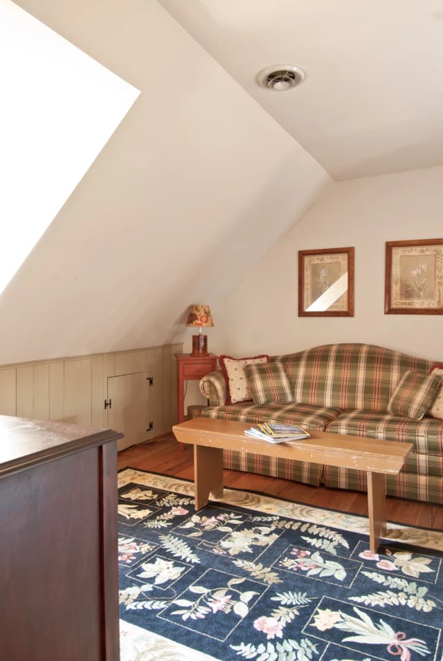 Cozy attic room with sloped ceiling, plaid couch, and floral rug. A wooden table holds magazines. Warm tones and artwork create a welcoming vibe.