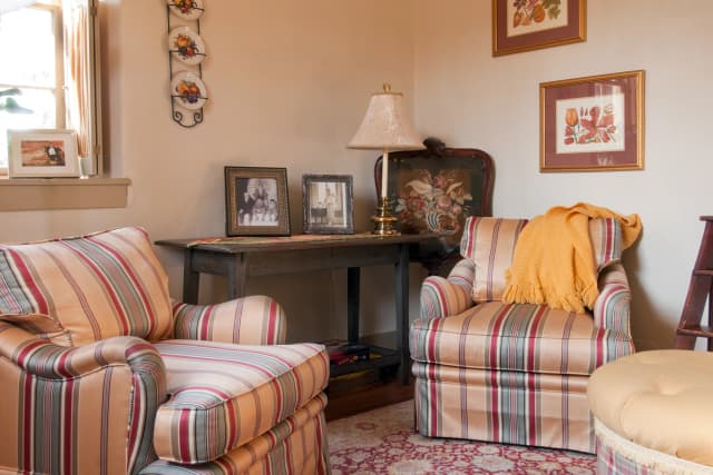 Homey living room with striped armchairs and yellow throw. A wooden table holds two framed photos and a lamp. Floral art hangs on beige walls. Warm and inviting.