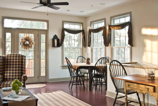Bright sunroom with dark wooden floors, a checkered armchair, and a rustic dining table with black chairs. French doors and windows draped with dark curtains add warmth.