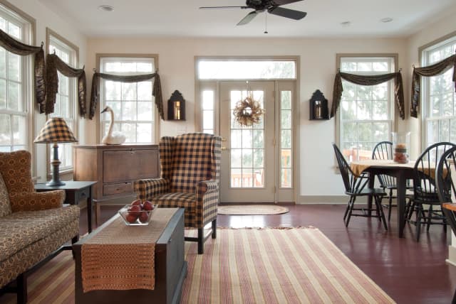 A cozy living room features a checkered armchair, patterned sofa, and rustic wooden furniture against large windows. Warm tones and soft natural light create a welcoming ambiance.