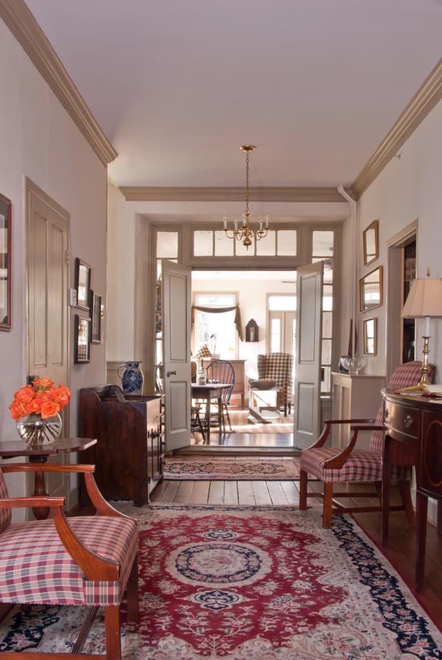 Elegant hallway with red patterned rug, plaid chairs, and a small table with orange flowers. Chandelier and vintage furniture create a warm, classic ambiance.