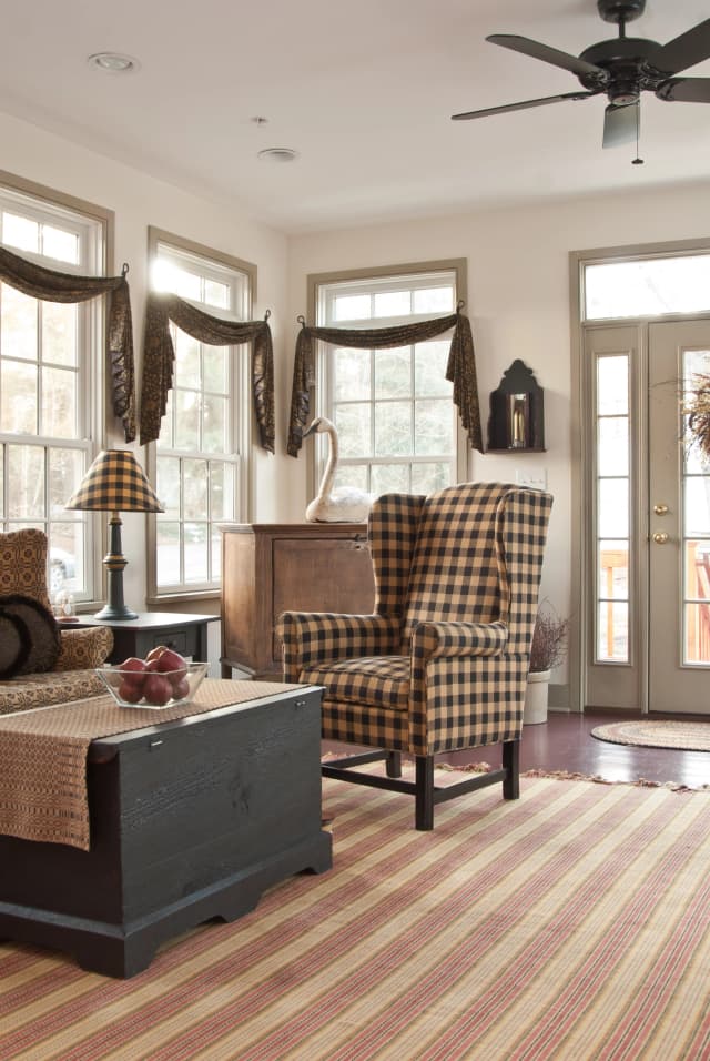 Cozy living room with checkered wingback chair, striped rug, and a wooden chest. Sunlight streams through large windows, creating a warm, inviting atmosphere.