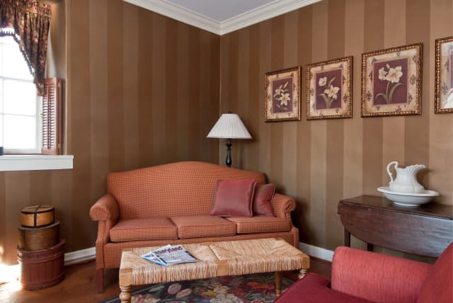 Cozy room with brown striped walls, red sofa, and matching armchair. Floral art, table with pitcher, baskets by window, warm, inviting ambiance.