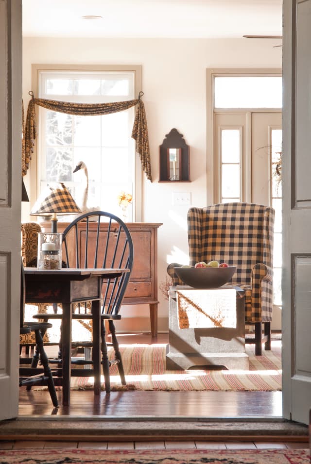 Cozy living room with warm sunlight, featuring a checkered armchair, wooden table, navy chair, and rustic decor, creating a welcoming atmosphere.