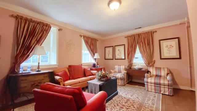 Cozy living room with pink walls, featuring a red sofa, striped armchairs, and floral decor. Warm lighting creates an inviting, relaxed atmosphere.
