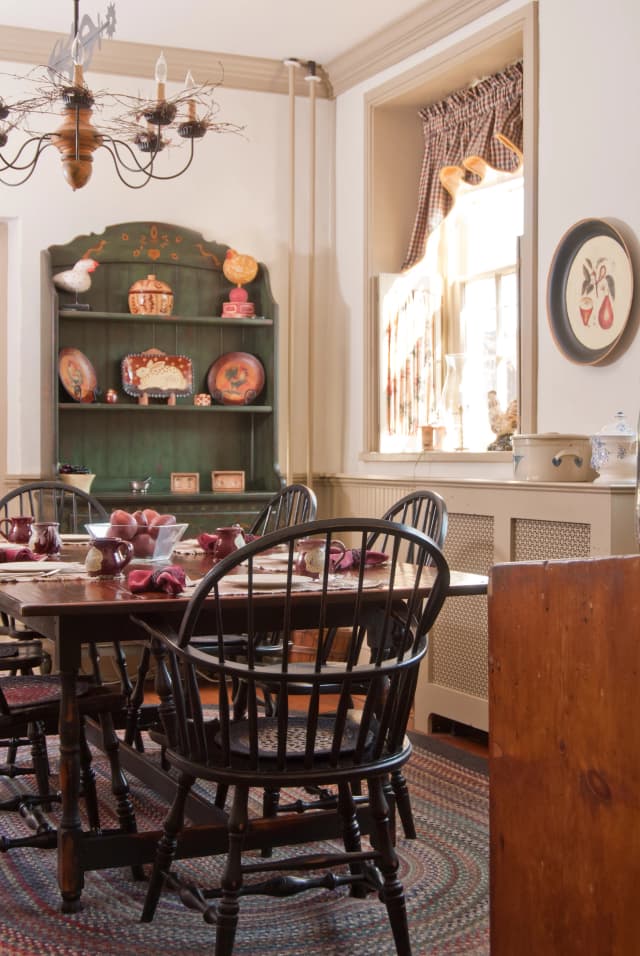 A dining room with a large wooden table and brown spindle-back chairs. A rustic green hutch with ceramics and roosters, and a softly lit window create a warm, inviting atmosphere. A braided rug and vintage chandelier add charm.
