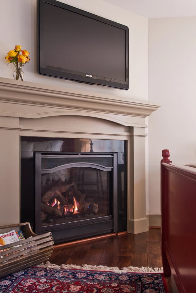 Cozy room with a lit fireplace beneath a mounted TV. A vase with yellow flowers decorates the mantel. A rack of magazines and a red chair complete the warm setting.