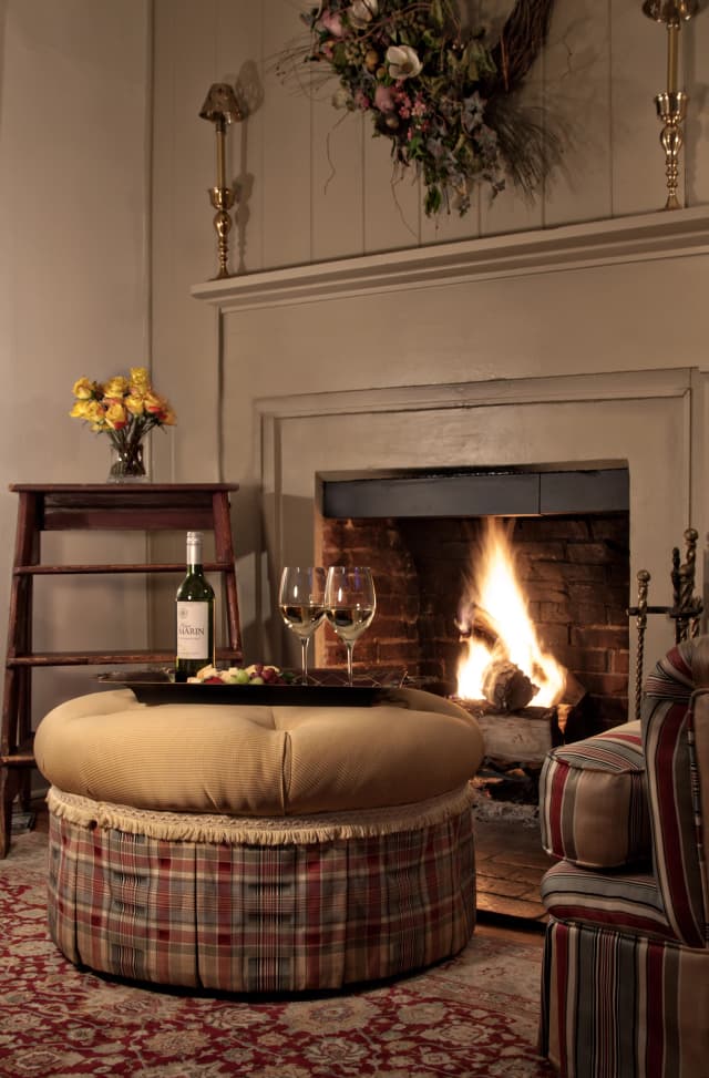 cozy living room with a lit fireplace, surrounded by a plaid ottoman holding wine and glasses. A bouquet of flowers and wreath add warmth.