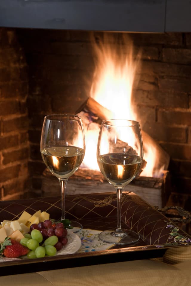 Two glasses of white wine, a plate of grapes, strawberries, and cheese sit on a tray in front of a cozy, roaring fireplace, creating a warm, intimate ambiance.