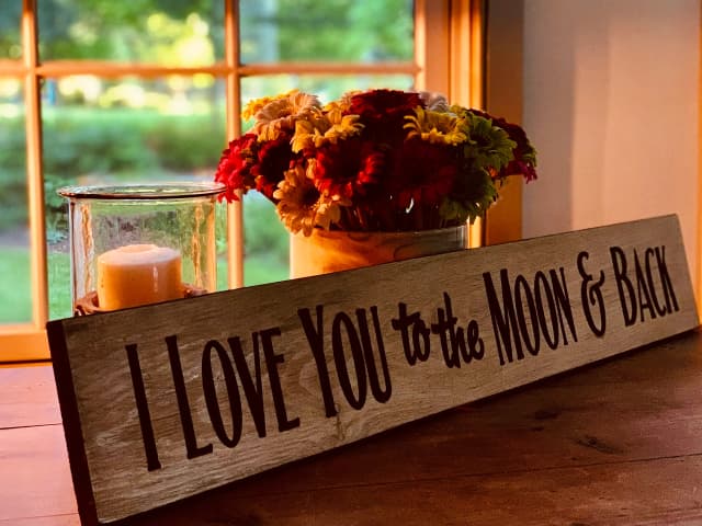 A wooden sign reading 'I Love You to the Moon & Back' sits on a table beside a lit candle and a vase of flowers, with a soft, warm light from a window.
