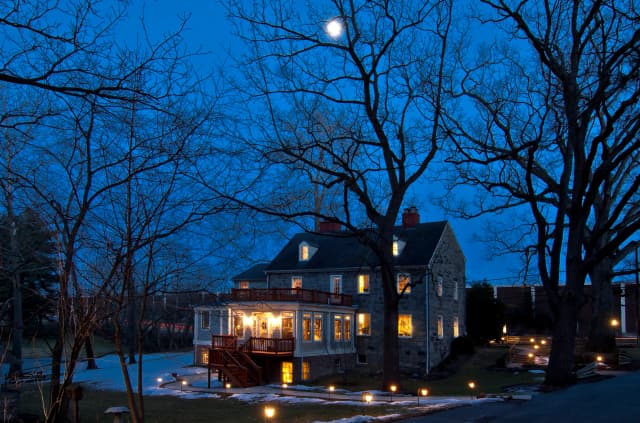 Cozy stone house at dusk, warmly lit, surrounded by bare trees under a bright moonlit sky. Pathway lights create a serene, welcoming atmosphere.