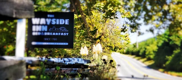 A blurred sign for 'The Wayside Inn Bed & Breakfast' is in the foreground, with trees and a winding road stretching into the sunny distance.